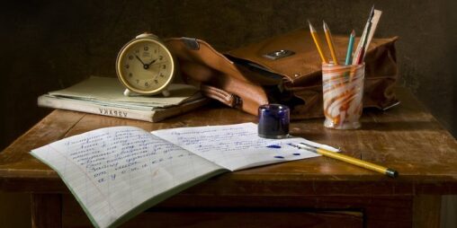 A still-life photo of a notebook, clock, ink jar, a bag, and pencils in a cup all on a desk